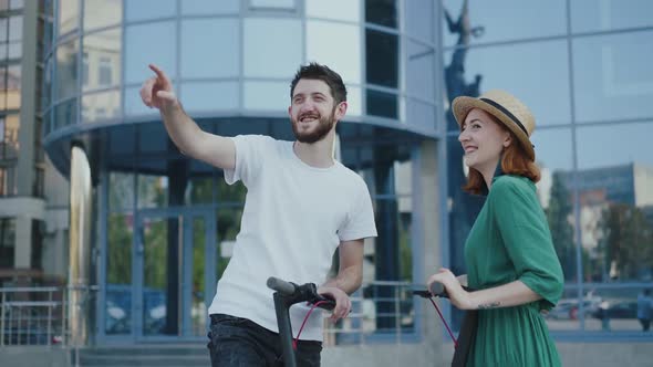 Happy Man Points Finger for Lovely Girl and They Smile When Standing on Scooters