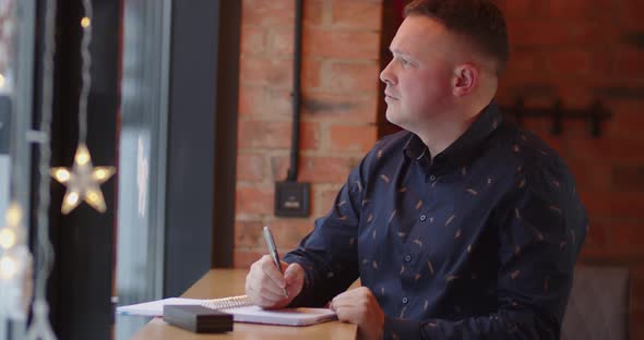 The Man Makes His Schedule. Man Writes Text In A Notebook, Sitting In A Cafe Looking Out The Window