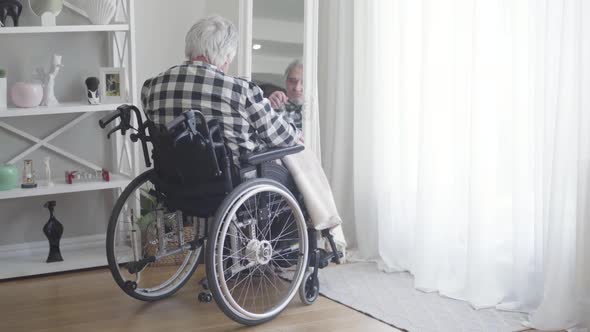 Back View of Mature Caucasian Man in Wheelchair Looking at His Reflection in Mirror, Sad Elderly
