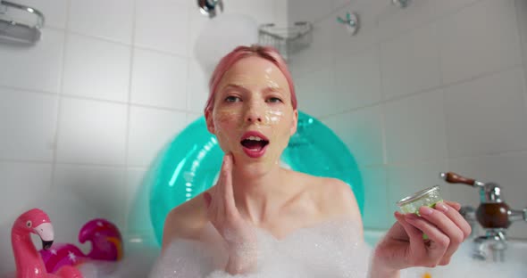 Cheerful Woman with Pink Hair Sits in a Bath in a Cloud of Foam and Makes a Cosmetic Mask