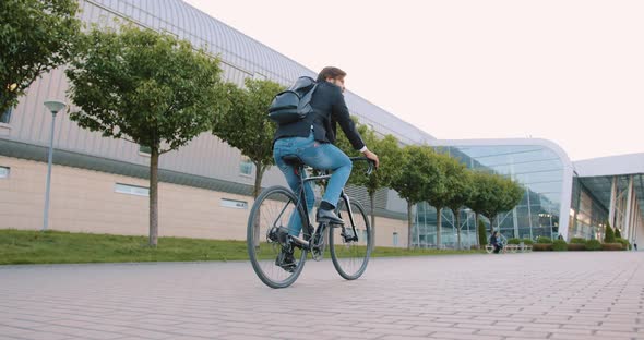 Guy Riding a Bicycle to His Work on Cobbled Path Near Modern Big Bulding with Glass Windows