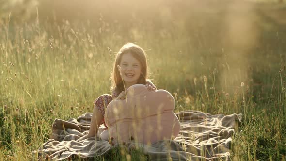 Little Cute Girl with Flowing Hair in a Summer Dress Sitting on the Grass and Playing with a Pink