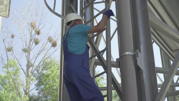 Side View of Factory Worker Climbing Up the Ladder on Enormous Stainless Steel Cement Storage Silo