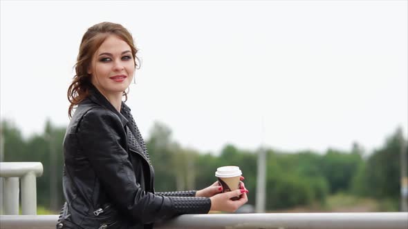 Romantic Woman in a Leather Jacket and with a Coffee in Her Hands Looks Away