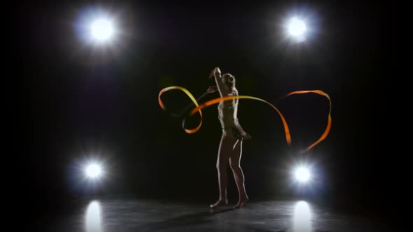 Gymnast with the Ribbon in His Hands Doing Acrobatic Moves. Black Background. Light Rear