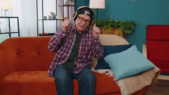 Overjoyed Senior Man in Wireless Headphones Dancing Singing on Cozy Couch in Living Room at Home