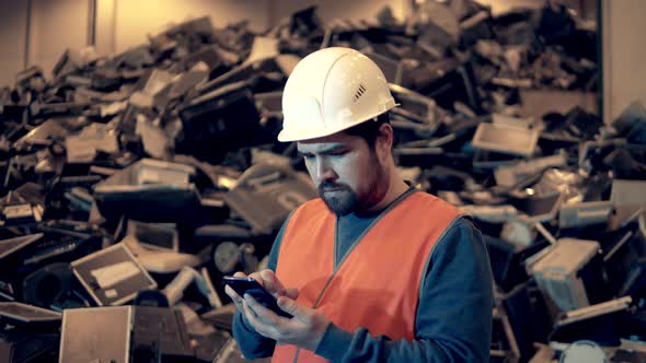 Garbage, Plastic Recycling Factory. Landfill Worker in a Hardhat Is Operating a Smartphone