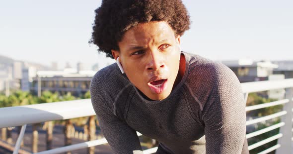 Fit african american man exercising outdoors in city, with wireless earphones, resting on footbridge