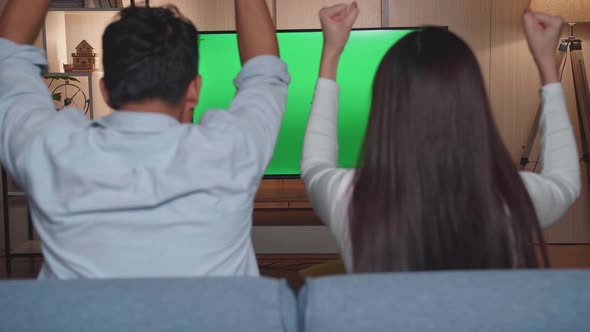 Back View Of Excited Asian Couple Watching Tv With Mock Up Green Screen Together Sitting On A Sofa