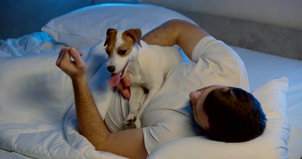 a Man Is Lying on a Sofa with White Bed Linen with His Dog. the Guy Is Stroking the Animal's Neck