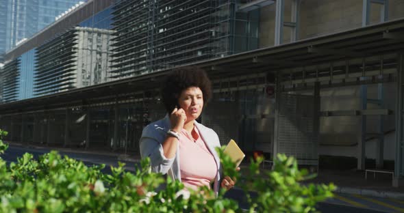 Plus size biracial woman talking on smartphone and holding notebooks in city