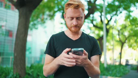 Pensive red haired man texting by smartphone while sitting on the bench