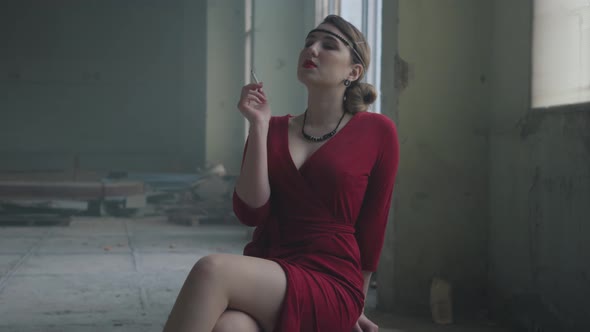 Elegant Woman in Red Elegant Dress Sitting on the Chair in the Abandoned Building Smoking Cigarette