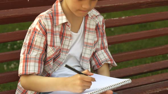 Little Boy Draws in His Notebook on the Bench