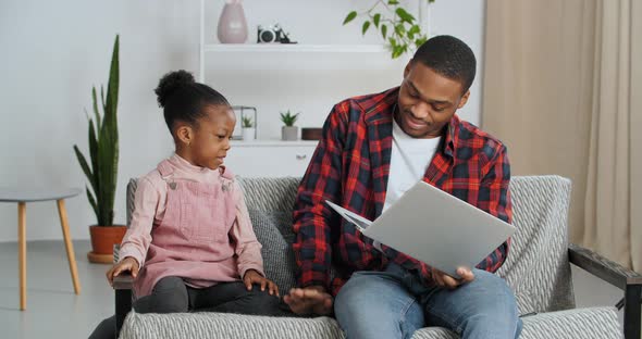 Young Father Afro American Guy Sits on Couch with Little Cute Girl His Daughter Teaches Child How to