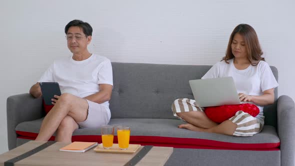 Asian senior couple sitting on a sofa and each of them looks at his own digital device.