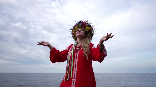 Smiling Ukrainian Woman in Red Dress and Head Wreath Raising Hands Looking Up at Cloudy Sky