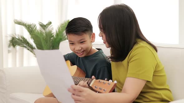 Asian mother and son sitting on the white sofa in the house son plays guitar, mother sings