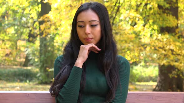 A Young Asian Woman Sits on A Bench in A Park and Thinks About Something Upsetting