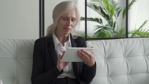 Elderly Woman Uses a Digital Tablet While Sitting on the Couch at Home