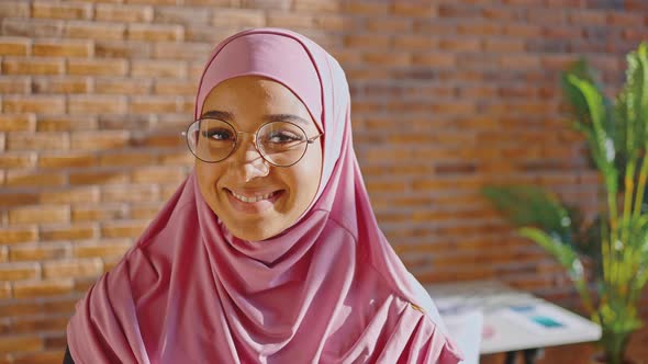 Muslim Woman in Pink Hijab and Glasses Smiling and Looking at the Camera Against the Background of a