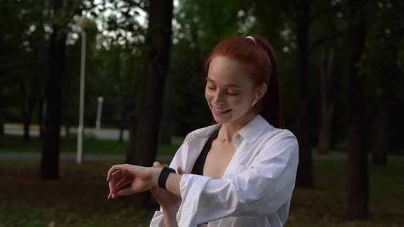 Smiling red-haired young woman is using application on smart watch outdoor in city street at evening