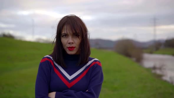 Portrait of a Brunette with Bright Lipstick on Her Lips and in a Blue Jumper Against a Green Meadow