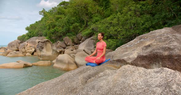 Woman Seats on Big Rock Against the Sea and Meditating She Wears Pink Sportswear and Uses Blue Mat