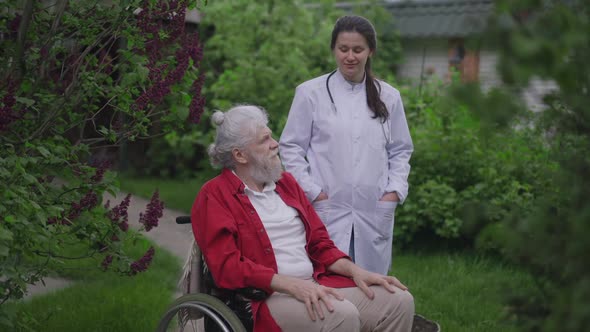 Confident Positive Caucasian Young Woman Talking with Handicapped Old Man in Wheelchair Standing in