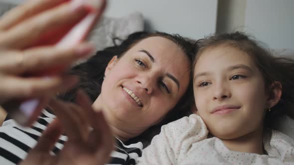 A Happy Woman and Her Daughter Lie Together on a Bed Take Selfies on a Smartphone or Communicate Via