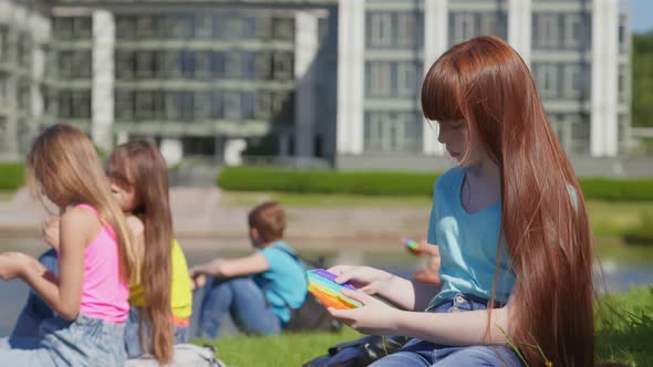 Preteen Caucasian Schoolgirl Sitting on Grass Outdoors Playing Pop It Toy