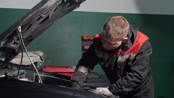 Young Worker in Uniform Examines to Fix Broken Automobile