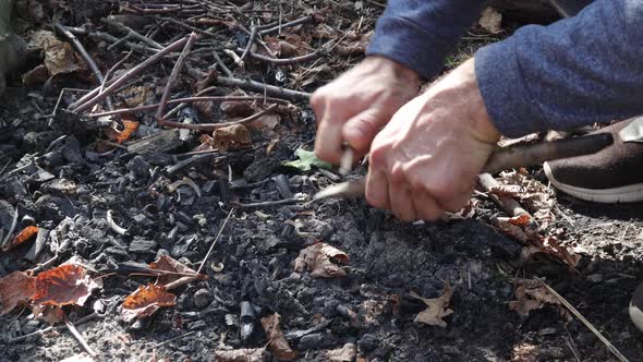 A man planes small branches for a fire with a knife