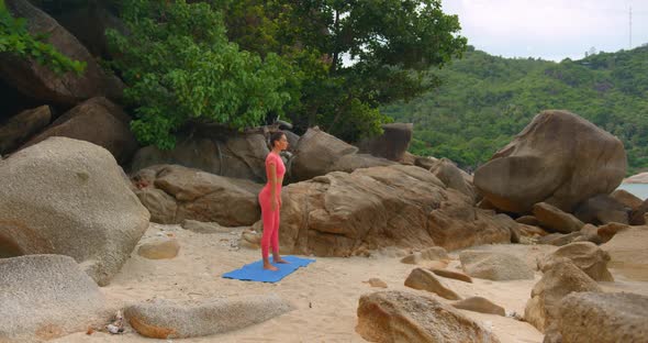 Sporty Girl in Pink Yoga Costume Making Workout on Blue Yoga Mat on the Beach Hidden Among Stones