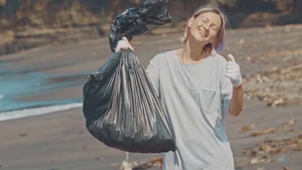 A Volunteer on a Black Ocean Beach with Plastic Trash in a Black Bag Shows a Hand Like Sign