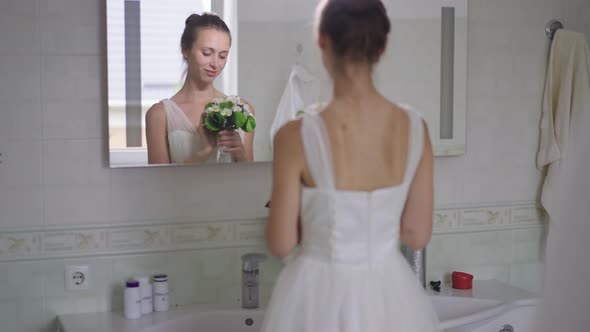 Romantic Happy Slim Young Bride Reflecting in Mirror in Bathroom Smelling Bouquet Smiling