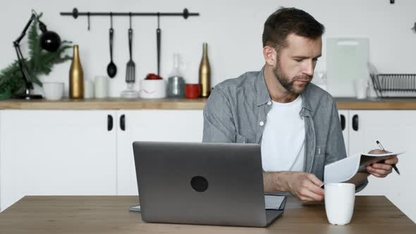 A Young Man Sits at a Table at Home and Working on a Laptop Writes down Notes