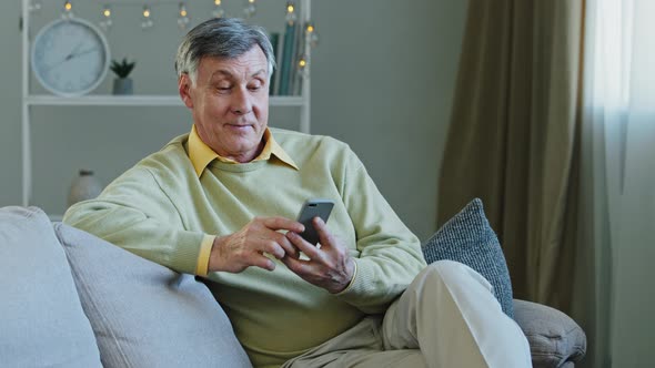 Happy Elderly Adult Man Sitting on Sofa Hold Gadget Studying App on Smartphone Makes Online Order