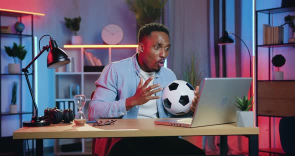 African American Man Holding Football Ball in His Hands During Revisioning Football Match on Laptop