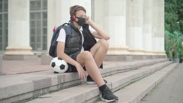 Wide Shot of Relaxed Schoolboy Sitting on School Stairs and Listening To Music in Headphones