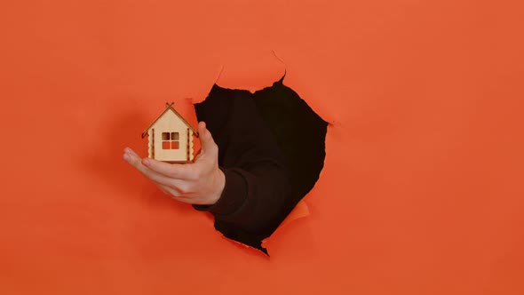 Man's Hand with Small Wooden House Sticking Out of Hole of Orange Background