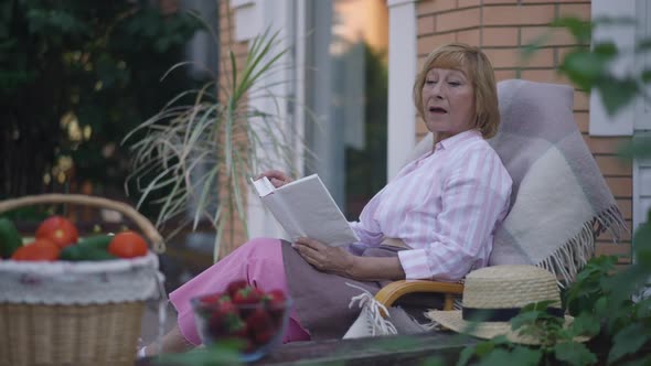 Side View Intelligent Absorbed Senior Woman Reading Book Sitting on Porch Outdoors on Spring Summer