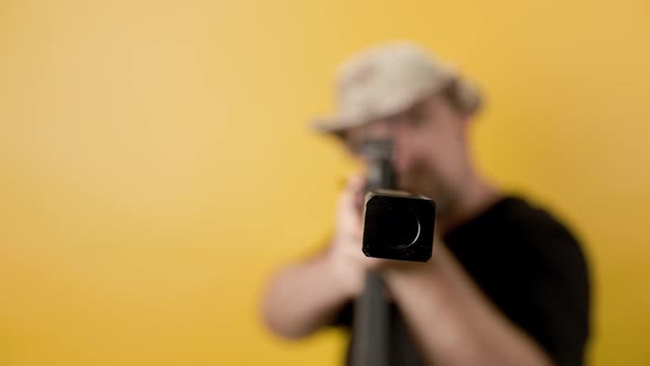 Middle aged man with rifle in black t-shirt and hat against yellow background.