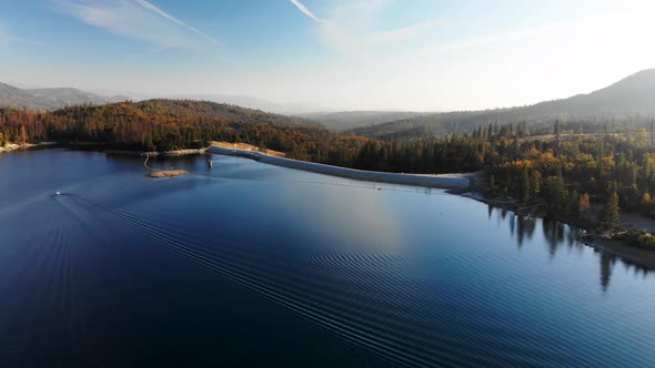 Aerial shot of a dam on a blue alpine lake surrounded by pine trees and mountains