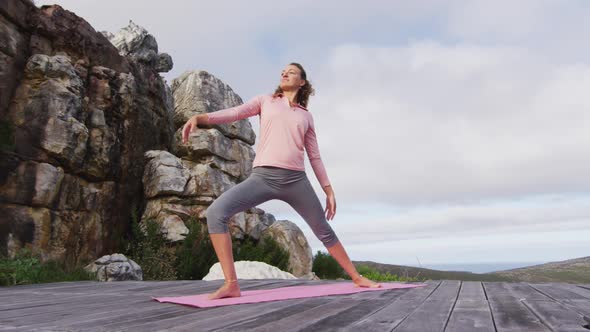 Caucasian woman practicing yoga outdoors, stretching standing on deck in rural mountainside setting