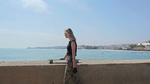 Pretty Young Girl with a Skateboard in the Hands Walking Along Seafront