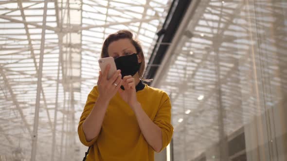 Busy Cheerful Girl with Protective Face Mask Walks Looking at Her Phone in in Shopping Center. Glass