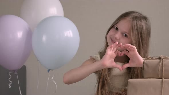 Cute Little Girl Making Heart with Her Hands and Smiling Surrounded By Air Balloons and Gifts