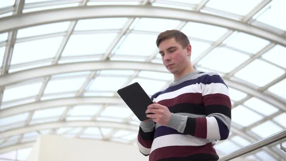 A Young Man Is Standing with a Tablet. A Man Uses a Tablet Against a Pan-glass Roof. A Busy Person