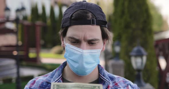 Man in Mask and Gloves Holds Last Dollar in Hands Looks at Camera with Sadness